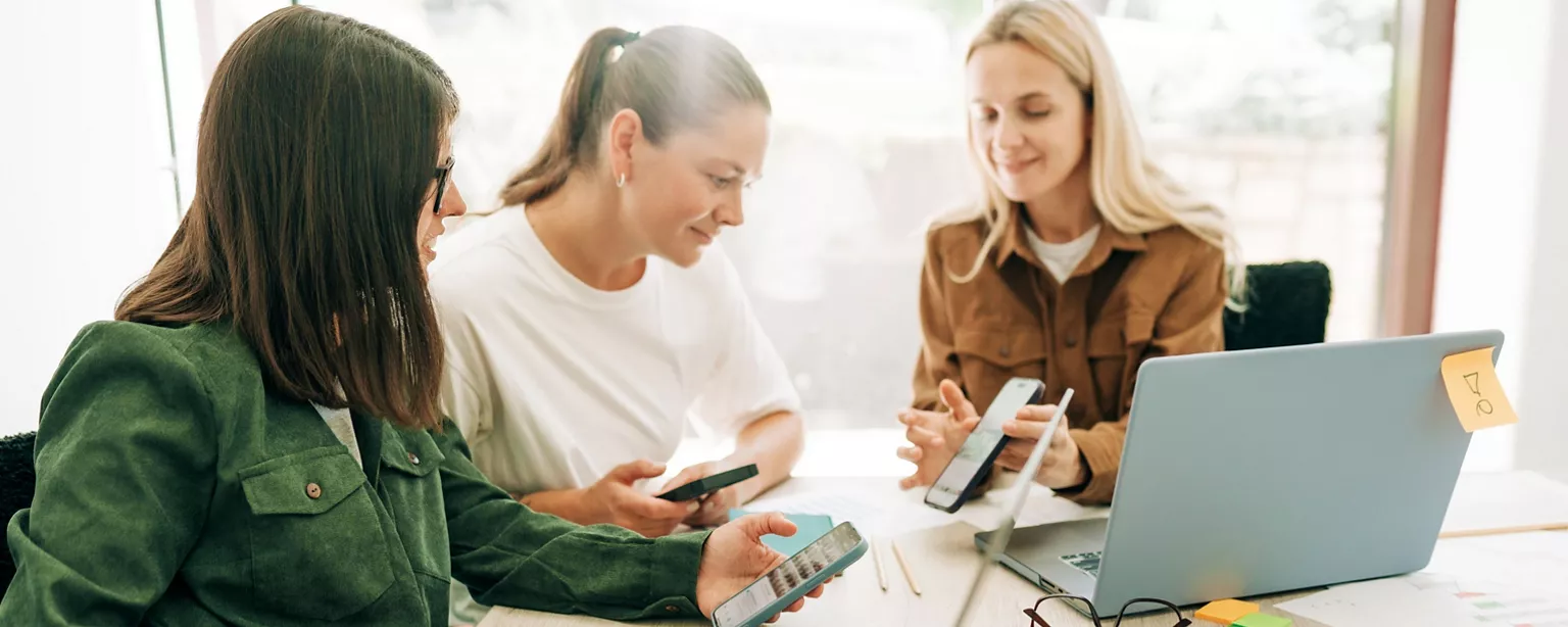 A photographic image of three women in an office setting looking at a laptop and their phones examining social media content.