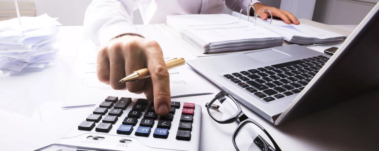 A man who is a tax accountant is working at his desk in front of an open binder, using a calculator and a laptop.