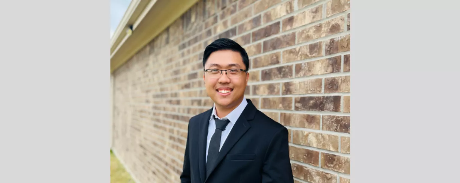 The Huan Hoang, Robert Half cybersecurity apprentice, smiles as he stands outside with a brick wall as a backdrop. 