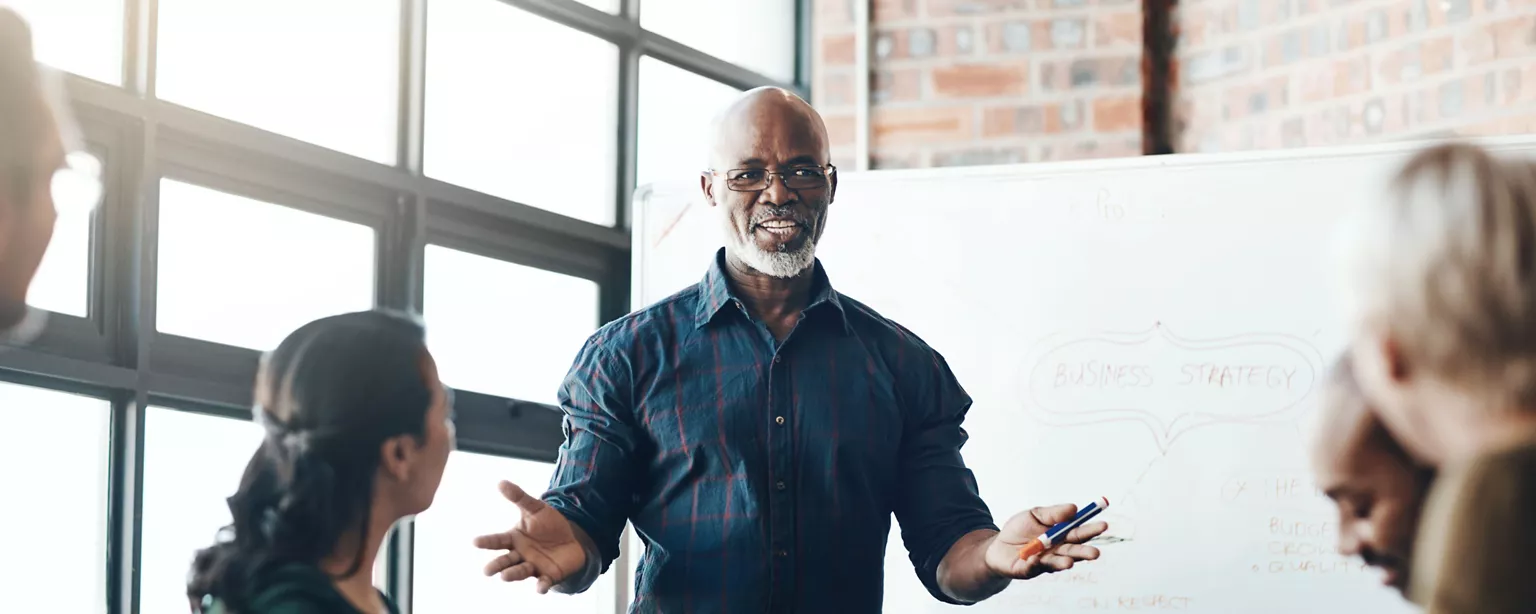 A man in a blue shirt, wearing glasses, stands in front of a white board, talking to a group of people sitting at a table.