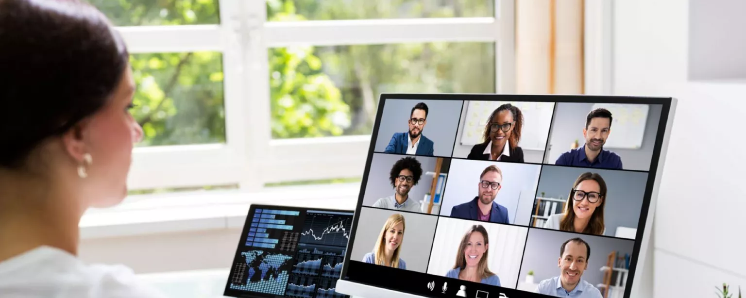 Woman in a white blouse sitting at a desk and typing on a keyboard in front of two computer screens, one showing financial charts and the other showing coworkers on a video call.