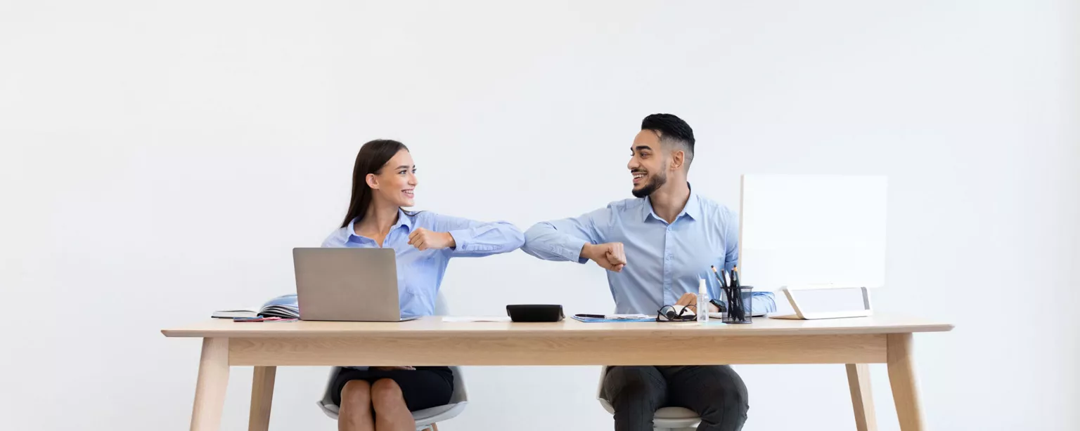 Smiling coworkers, a woman and man, sit next to each other at a wooden table and bump elbows.