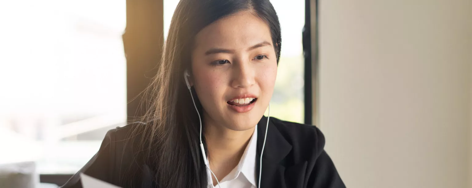 Young woman wearing a black blazer and headphones, using her laptop to participate in a video meeting as she gestures toward a document.