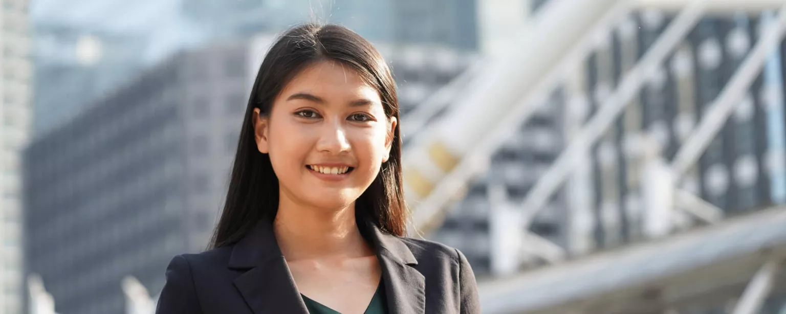 Young woman with long black hair in a black blazer and green shirt, standing against a backdrop of tall office buildings and smiling.