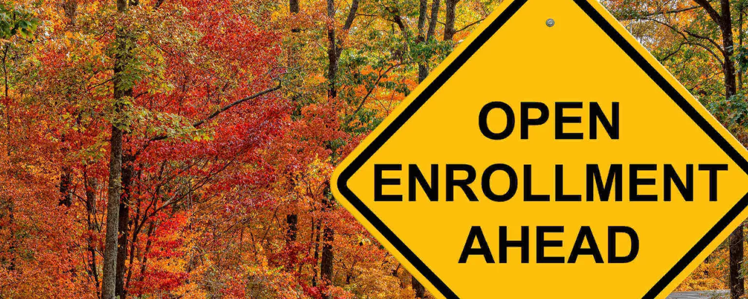 A yellow and black road sign along an empty country road in autumn that reads: "Open Enrollment Ahead."