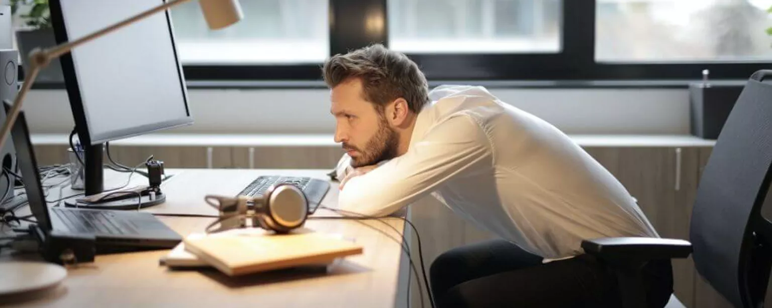 Ein Büromitarbeiter am Schreibtisch hat den Kopf müde auf die Arme gelegt und blickt auf seinen Bildschirm