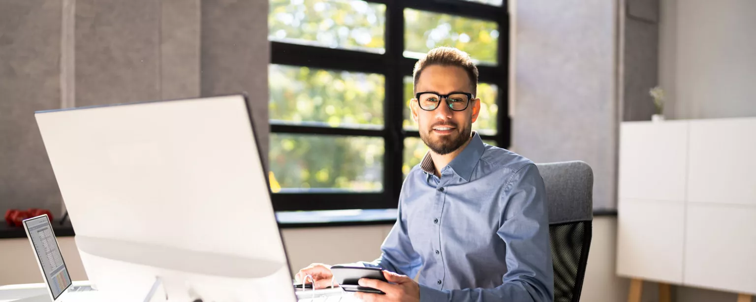 A young man with glasses, an accountant, sitting in his home office and smiling toward the camera while working on finances.