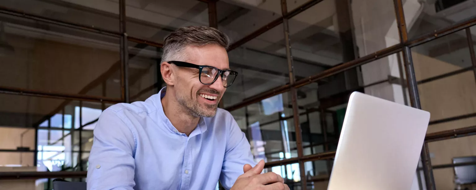 A smiling man in glasses and a blue shirt sits at his desk and engages in a video conference via his laptop.