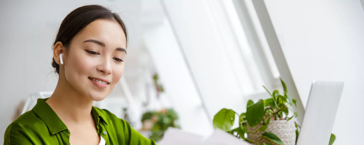 Young woman in a green blouse, working in a home office and smiling while reviewing documents in front of her laptop.