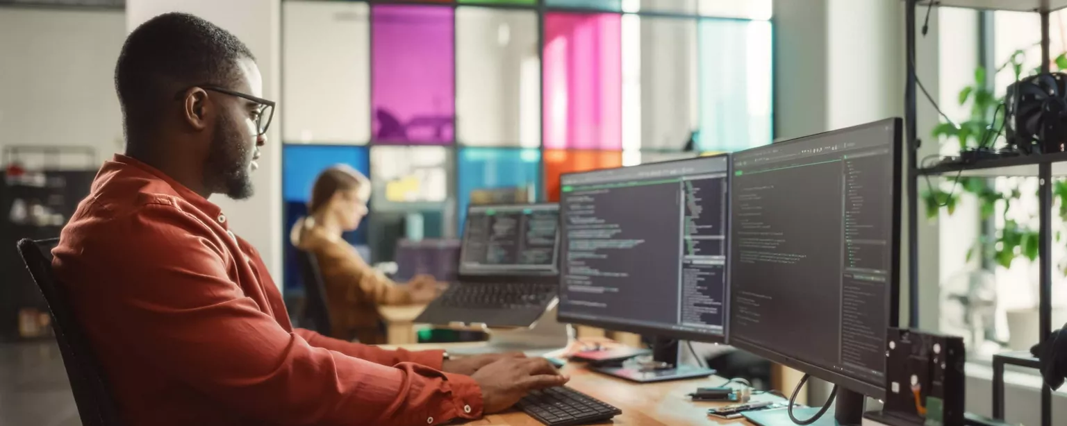 A developer who is working on code on a computer sits at a workstation in the modern-looking office of a tech startup.