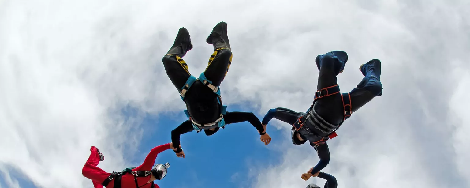 Skydivers in jumpsuits of various colors, forming a circle as they descend to the ground with a blue sky above them.