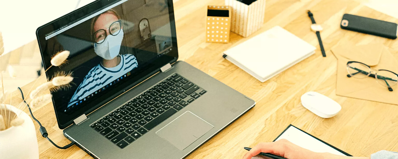 Eine Frau sitzt an einem Tisch mit einem Laptop, auf dem eine weitere Frau mit Mund-Nasen-Schutz zu sehen ist