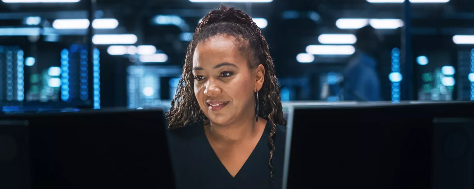 Young woman sitting in data center in front of two computer monitors, looking at one and getting ready to type on a computer keyboard.