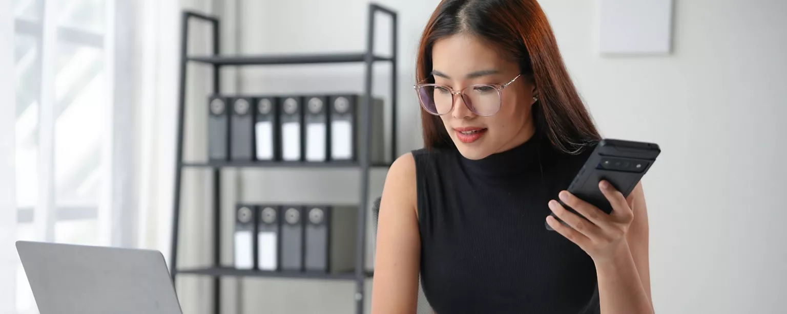 A controller works at her desk. She is holding her mobile phone while looking at her laptop.