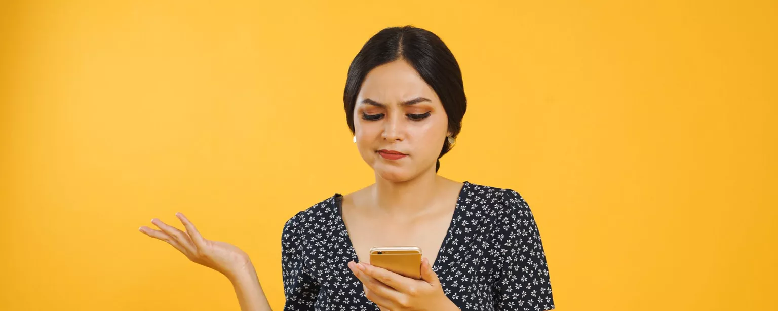 Young woman standing against a yellow background, staring at smart phone with a perplexed look and half-shrugging.