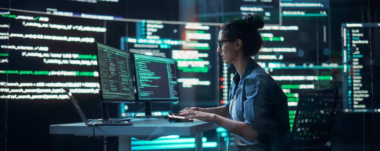 A woman who works in technology sits at a table in a security operations center looking at two computer screens and reviewing code..