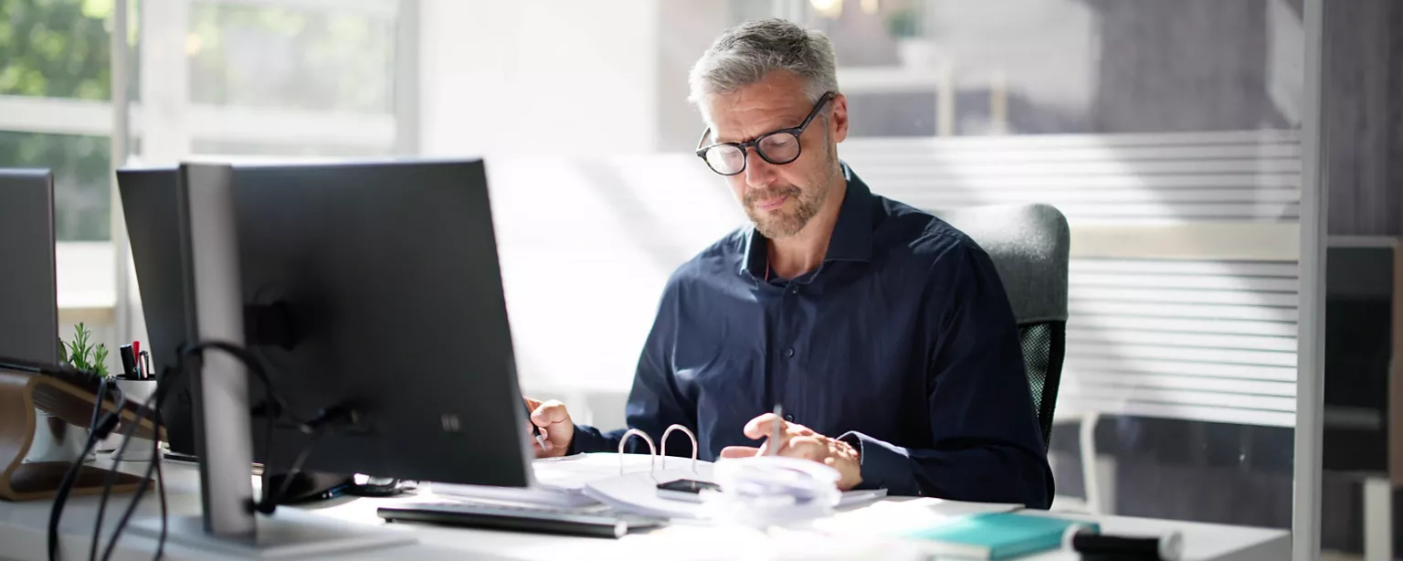 A finance consultant sits at a desk in front of a computer; he is concentrating on paperwork in a binder on his desktop.