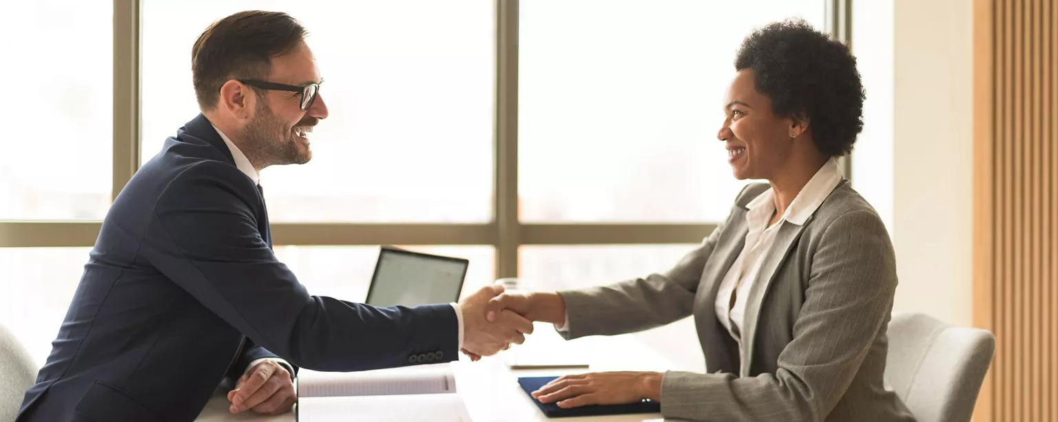 An executive recruiter and a candidate meet face to face; they are smiling and shaking hands.