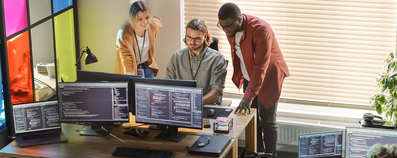 Three members of a technology team working at a startup company look at code on a computer.