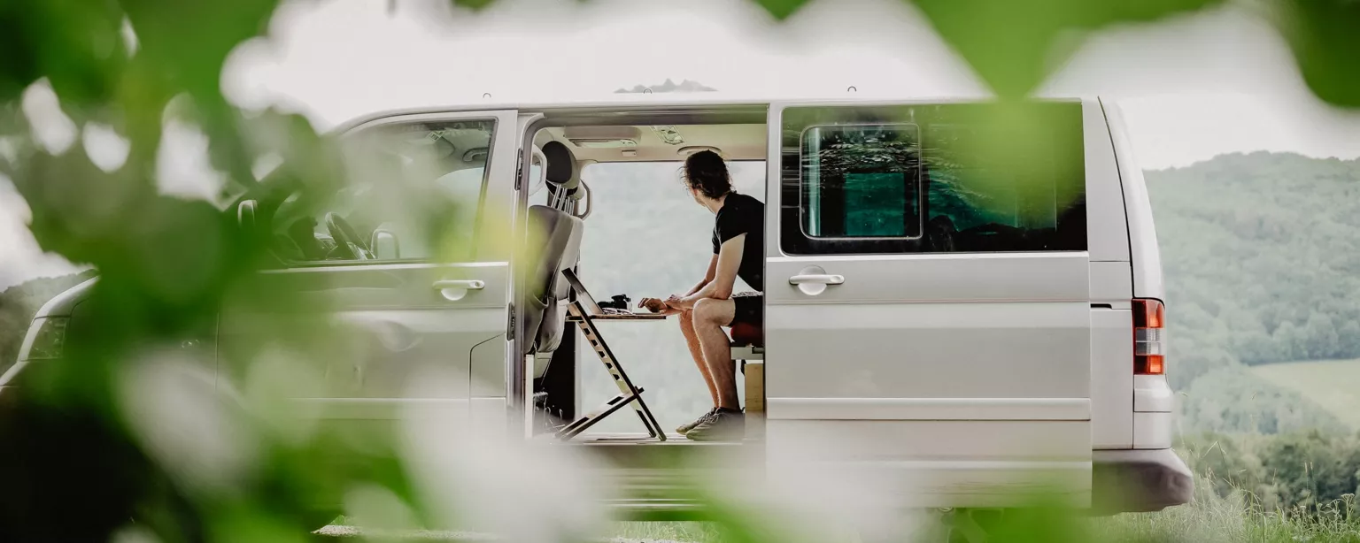 Ein Mann arbeitet in einem Van am Laptop und blickt in eine schöne Landschaft  