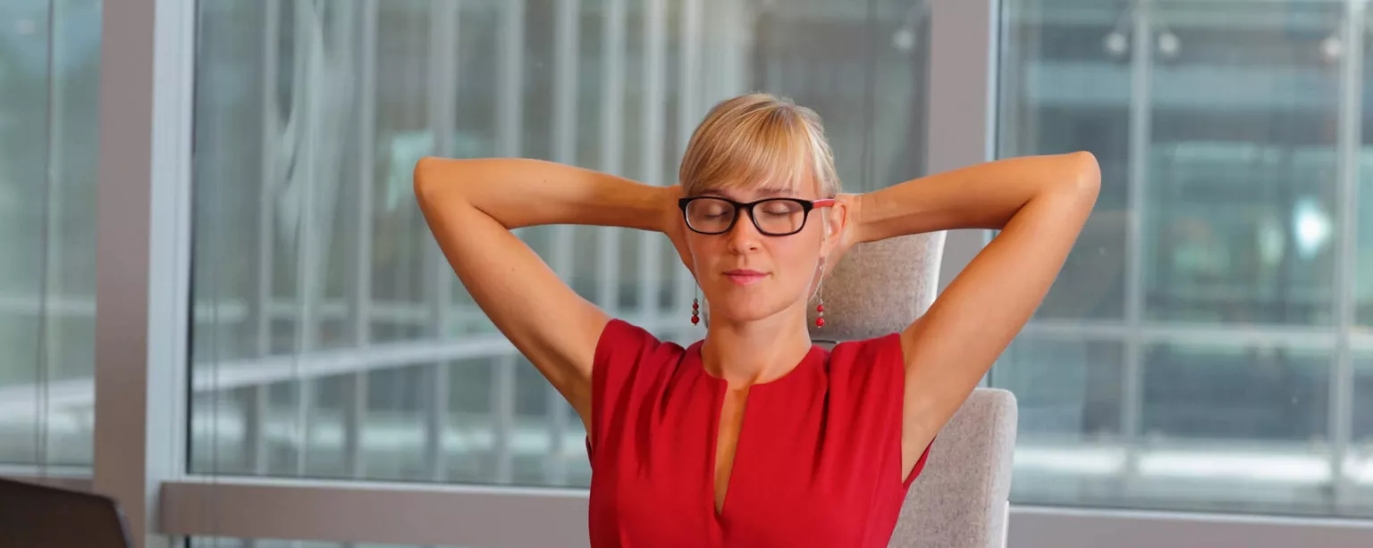 5 Workplace Wellness Trends to Watch — woman in red sits at office desk with hands behind head in relaxing stretch