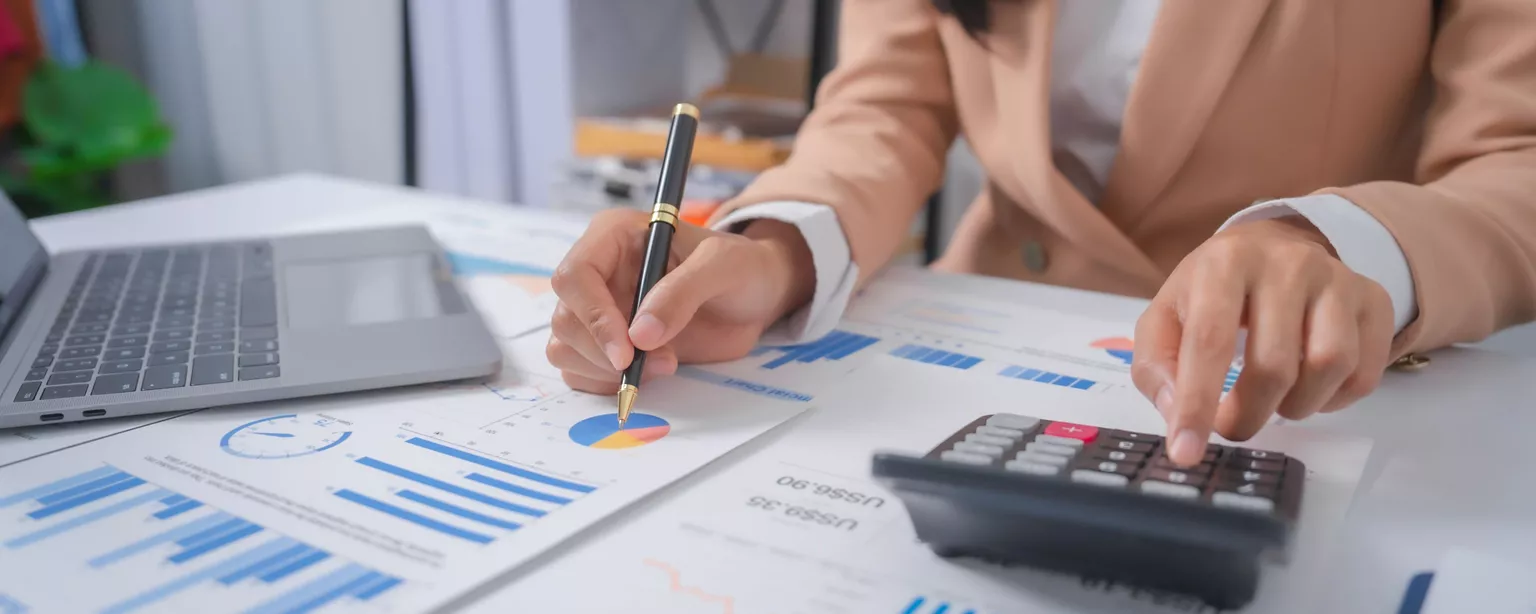 Finance professional reviewing financial reports and charts while using a calculator at a desk.