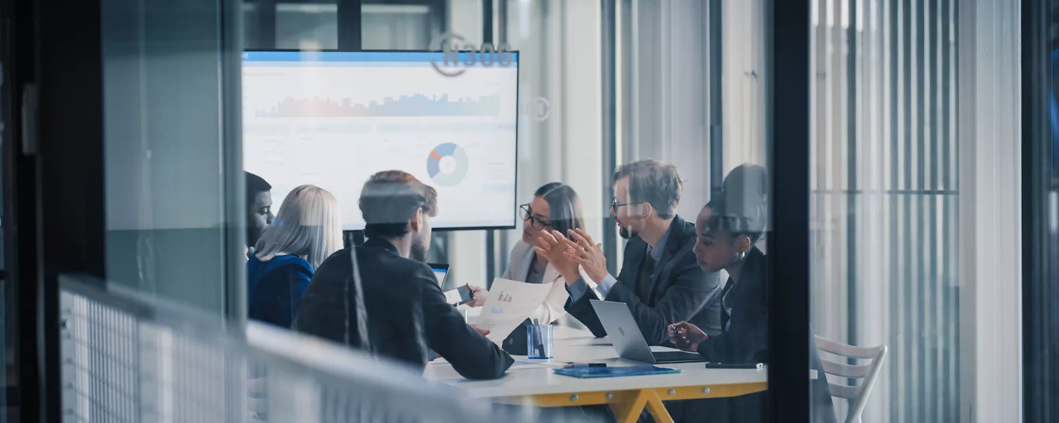 Finance professionals meeting in a conference room, reviewing reports and discussing data displayed on a screen.