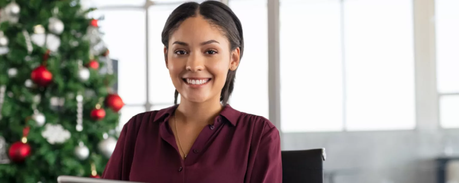 A young woman in a purple shirt holding a tablet computer and smiling while working in an office with a Christmas tree in the background.