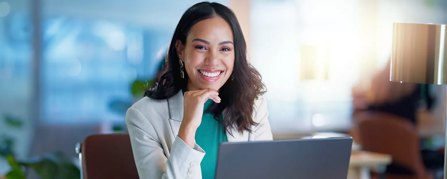 A professional woman smiles confidently while working on her laptop in an office.