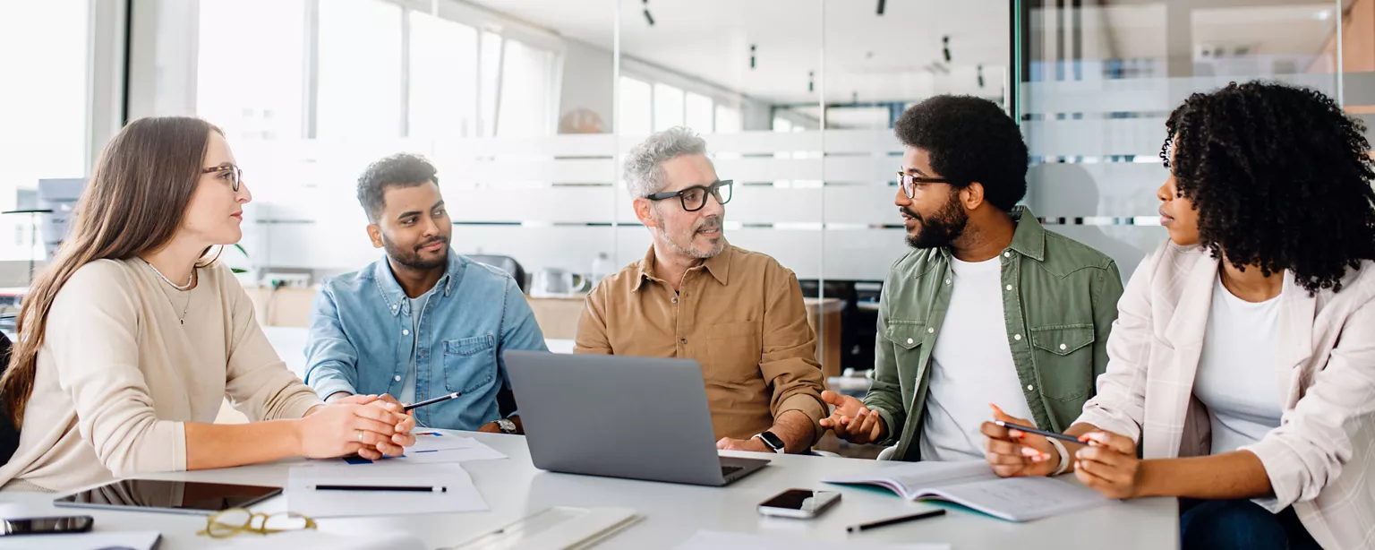 Multigenerational team of diverse coworkers gathered around a laptop in an office, discussing ideas during a meeting.