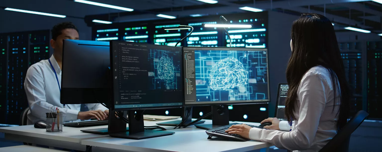 Two IT professionals work at computer stations displaying AI and automation graphics on dual monitors in a data center.
