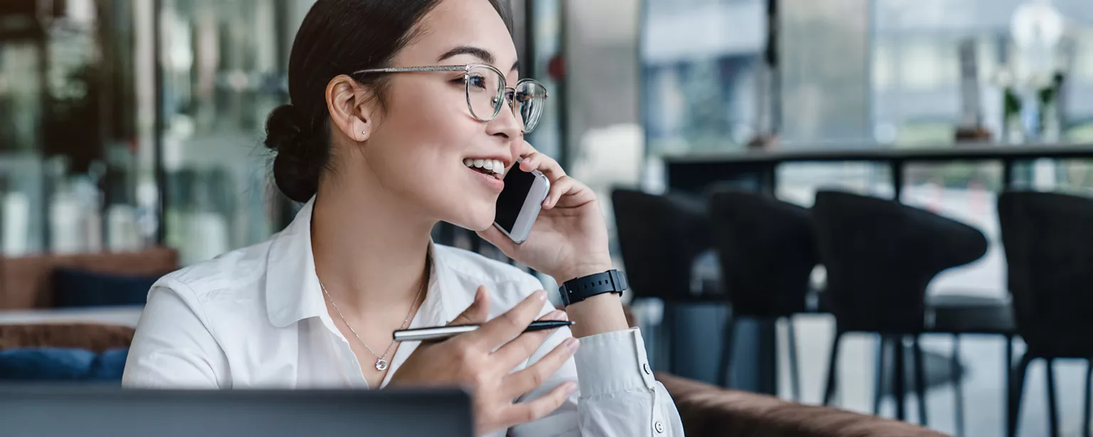 Enthusiastic-looking east Asian woman with black hair and glasses at a cafe speaking on a smartphone, with a pen in her right hand and a laptop in front of her.