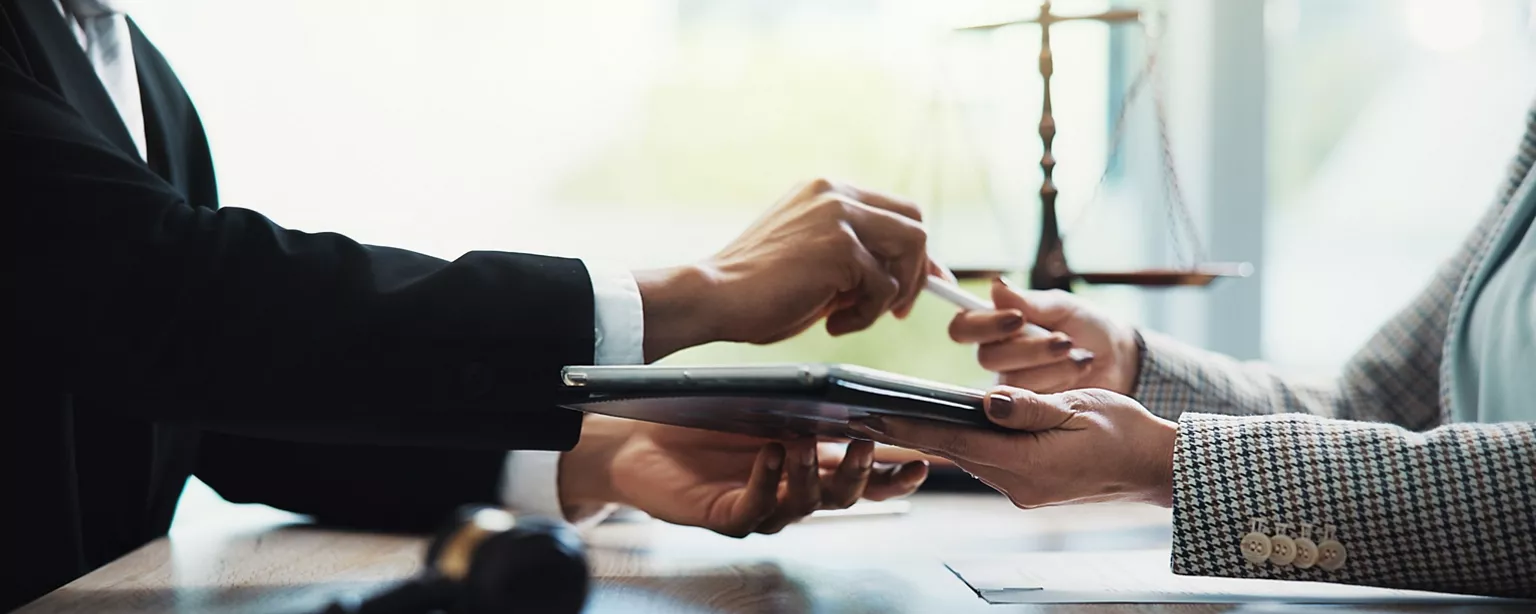 Two legal professionals review and exchange documents on a tablet in a modern office setting.