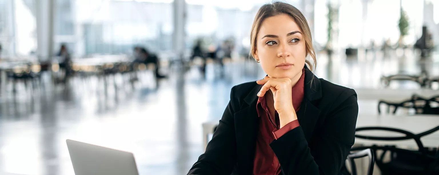 A thoughtful businesswoman sits at a desk with her hand on her chin, looking out into the distance in a bright, modern office setting.