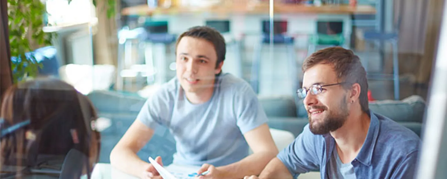 Two young businessmen listening to a person on the other side of a desk.