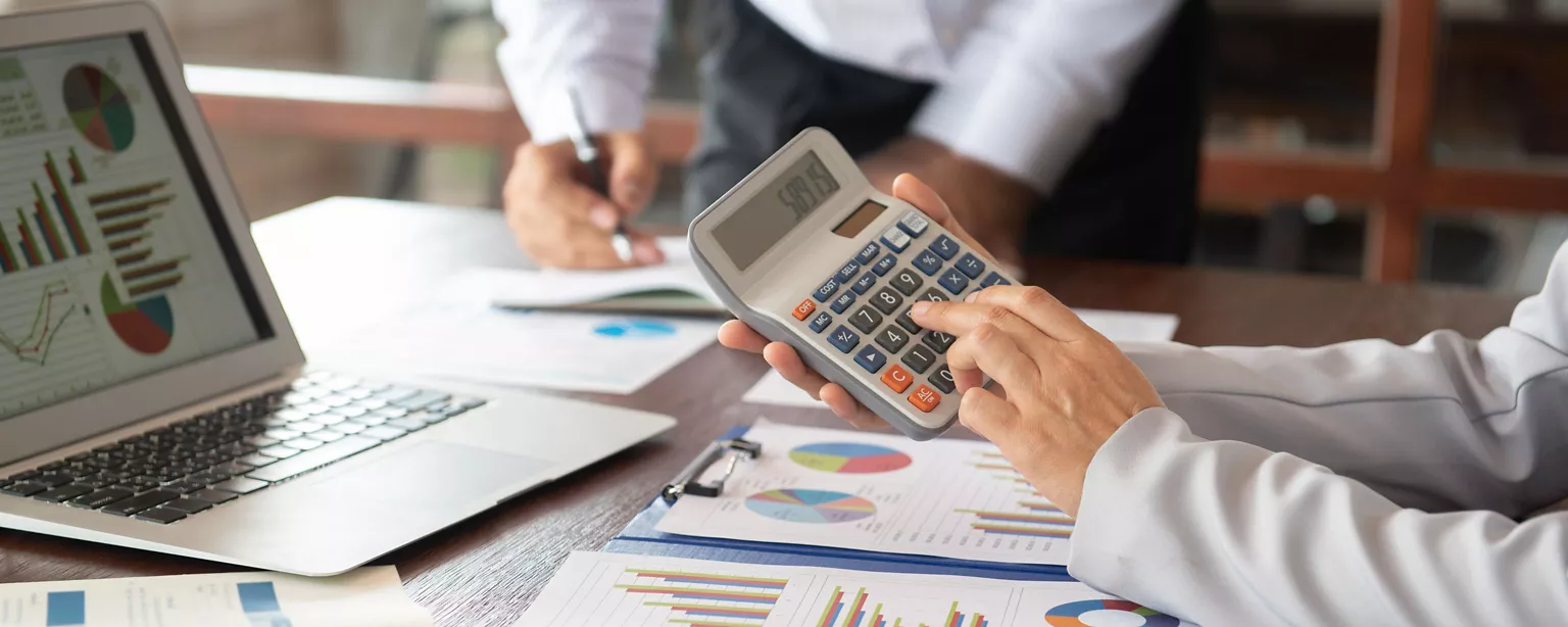Hands using a calculator with printed out charts beneath hands, and graphs displayed on the computer screen in the background, on an office desk.