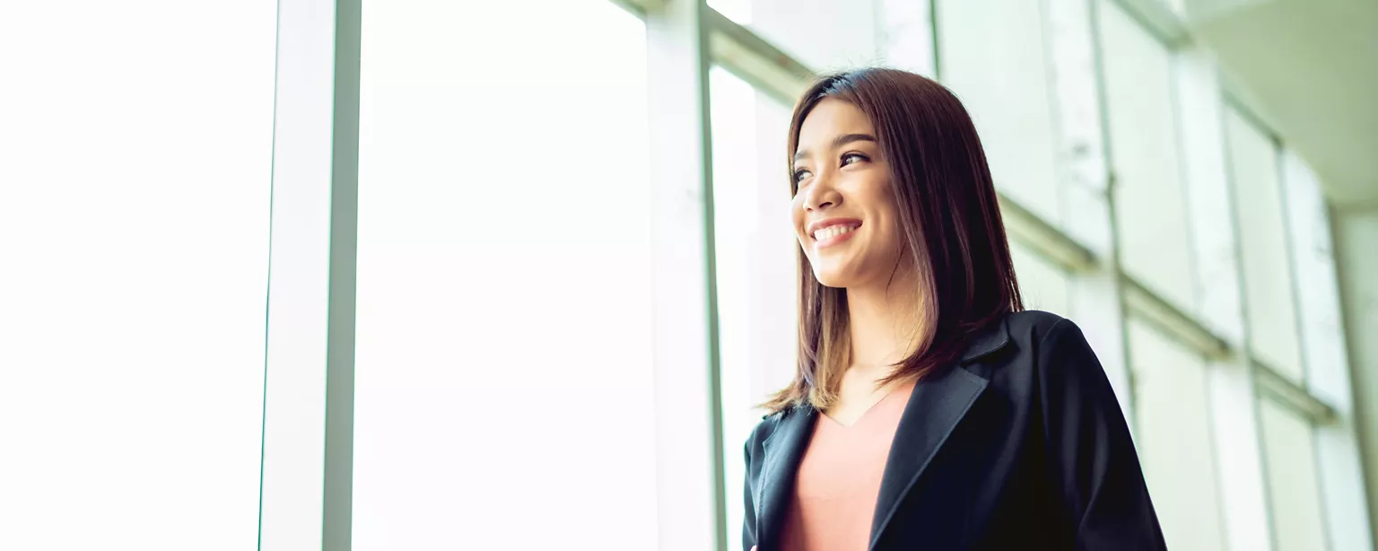 A smiling professional woman looks out a large window with confidence about a fresh start in her career.
