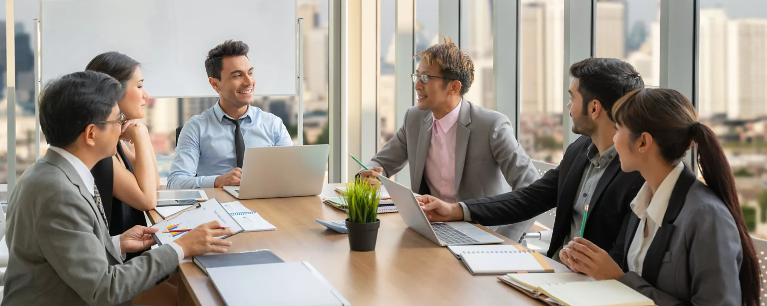 A group of professionals meets around a conference table, discussing work with laptops and notebooks in a bright office overlooking the city.