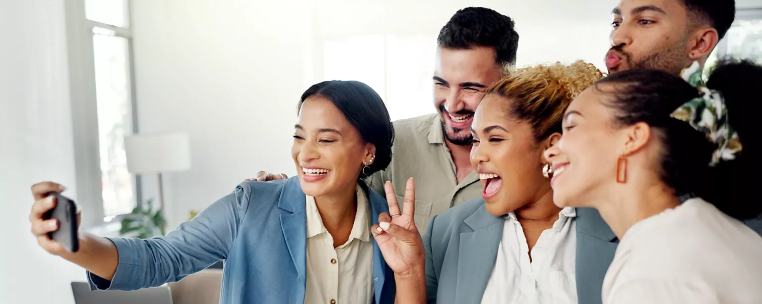 A group of diverse coworkers smiling and taking a selfie together in a bright office.