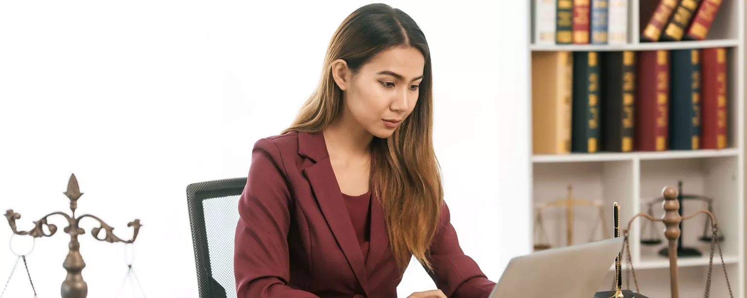 A focused young woman works on her laptop in a legal office, with shelves of legal books and scales of justice in the background.