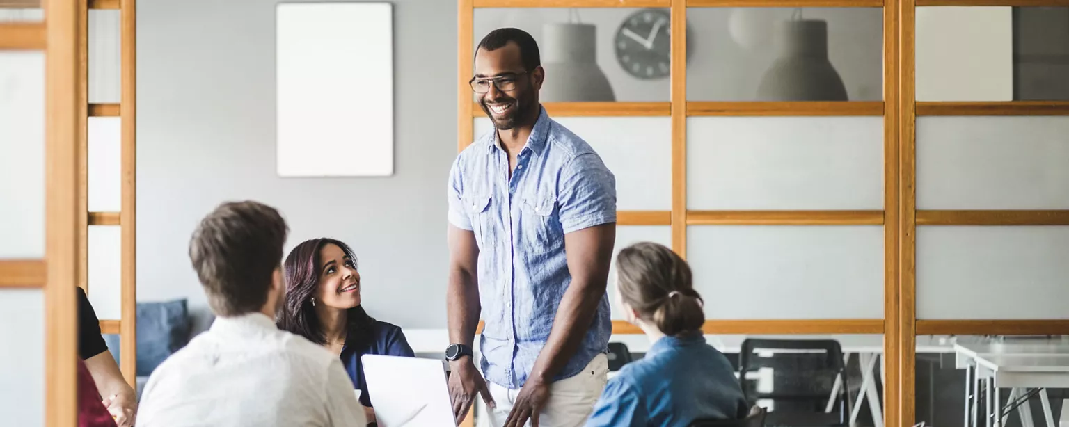 A team of colleagues smiles and listens as a man confidently speaks and stands during a casual office meeting.