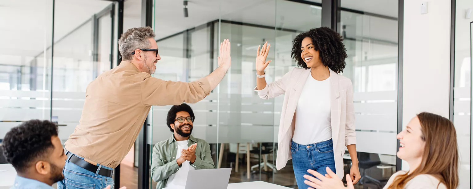 Coworkers celebrate success in a glass conference room as two colleagues share a high five while others applaud with smiles.