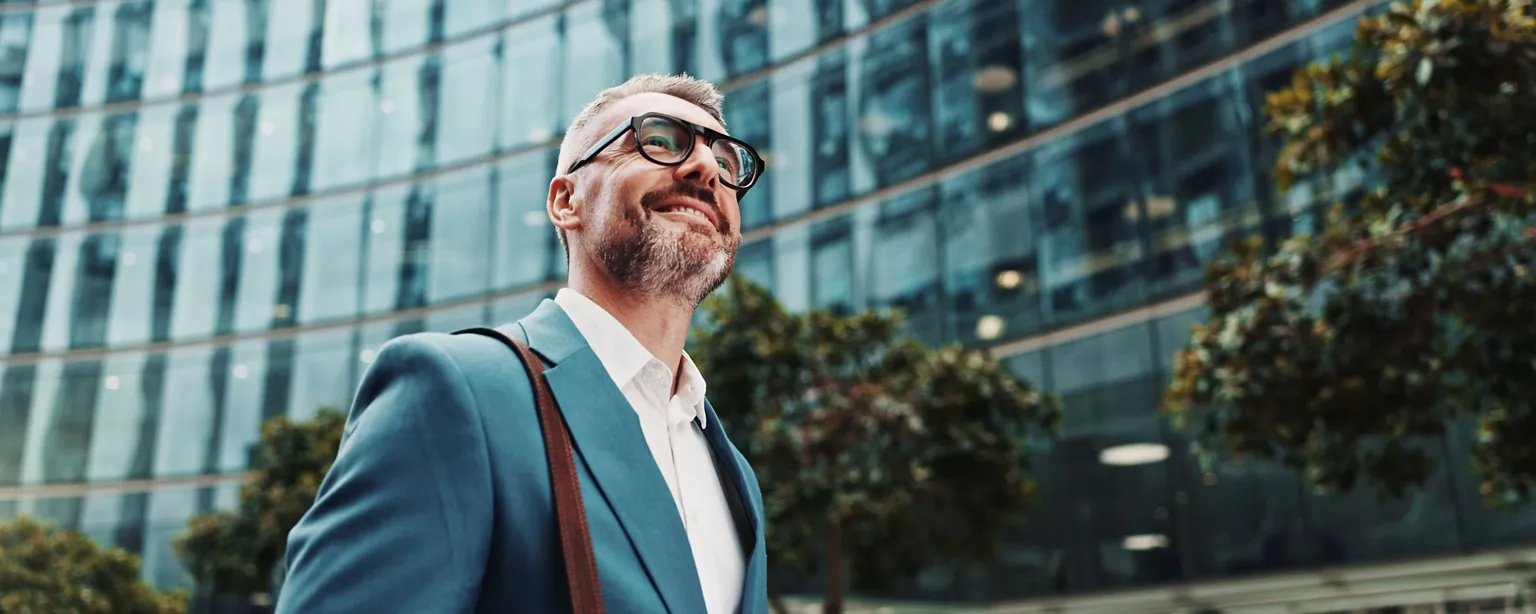 A confident business executive in a blue suit smiles while walking outside a glass office building.