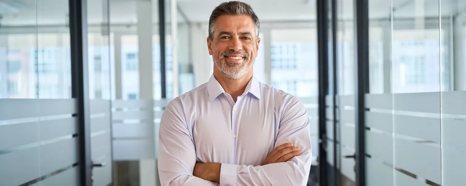 A man with arms crossed stands in an office smiling.