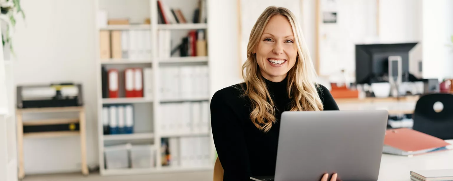 A smiling professional in her office working on customer success certifications with a laptop on her lap.