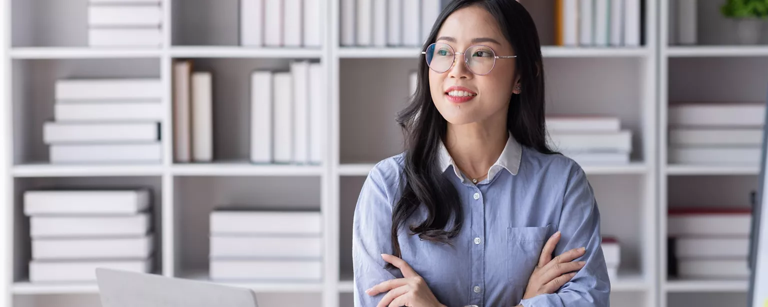 A confident professional woman in glasses sits at her desk with a laptop, notebook and calculator, smiling thoughtfully in an organized office space.