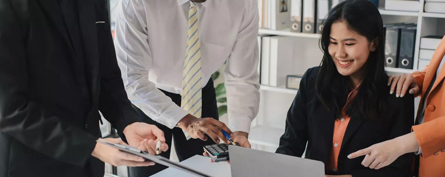 An accountant working at her desk smiles as several colleagues gather round to offer assistance.