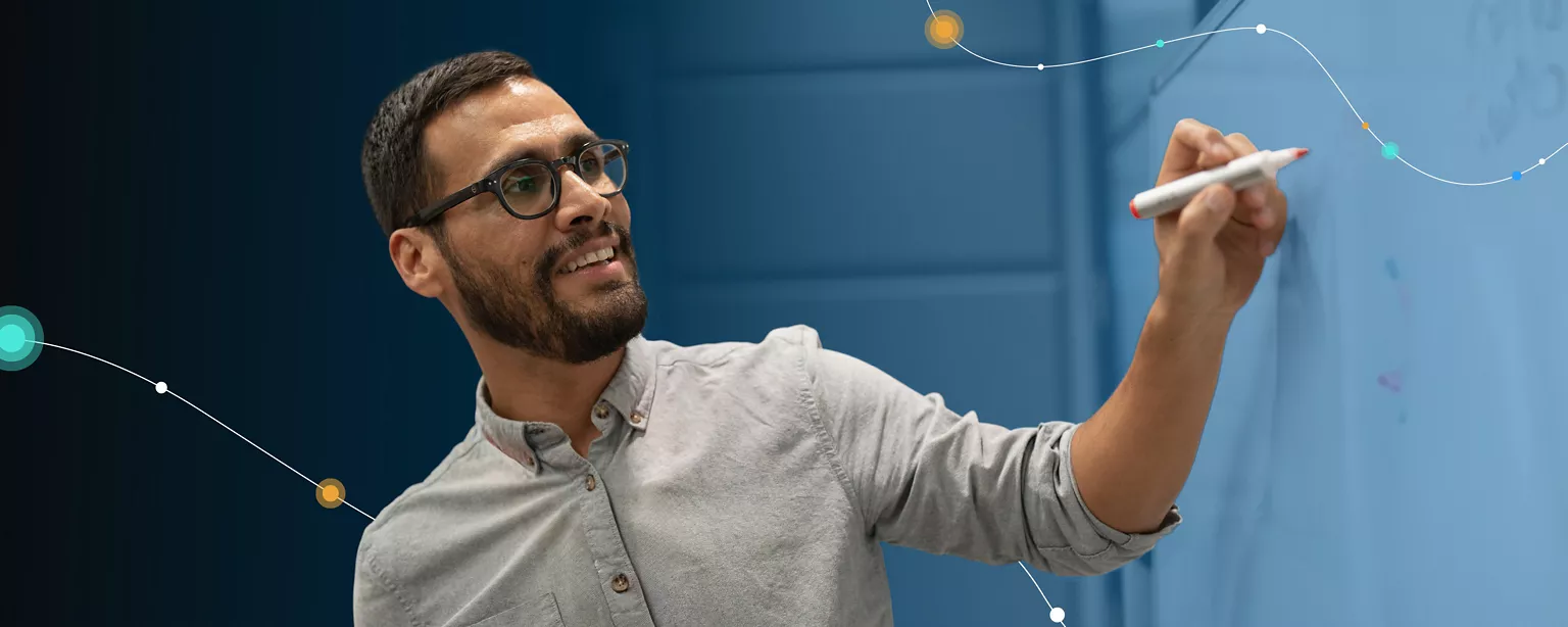 A man wearing glasses and a dress shirt smiles and stares at a blackboard while holding a marker in a marketing and creative environment.