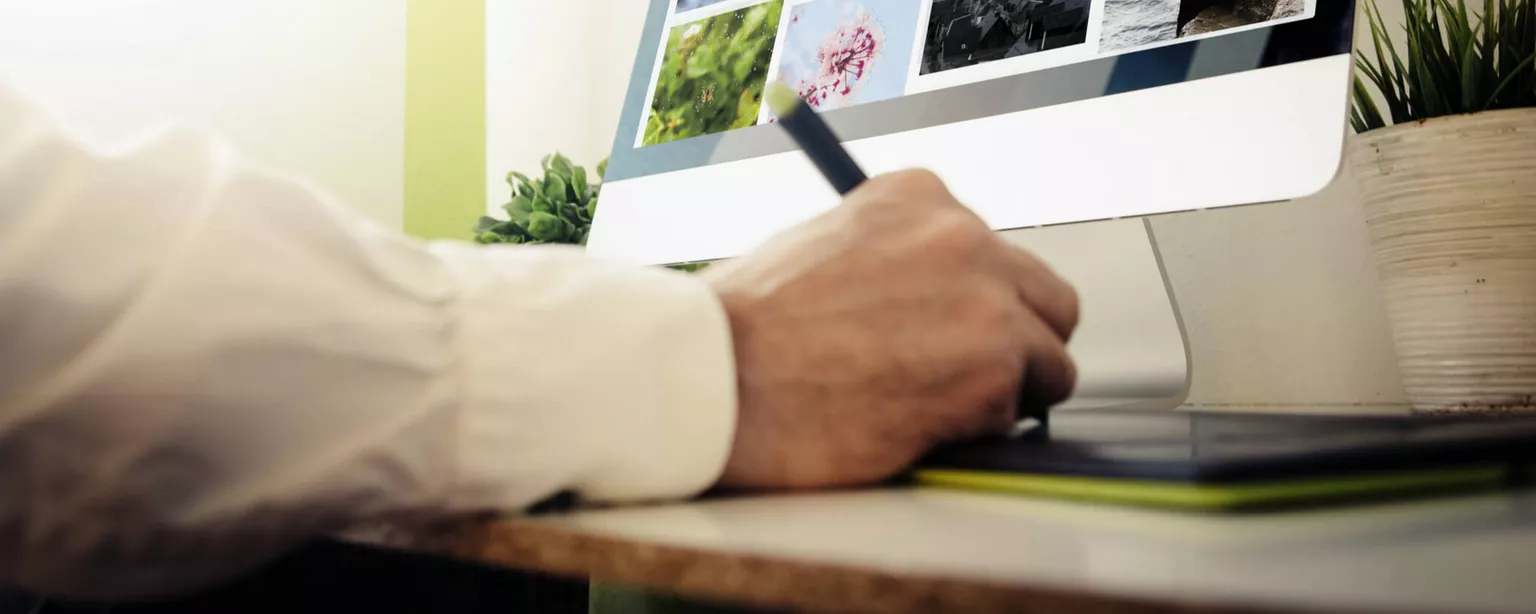 A hand in the foreground uses a drawing pad as a computer monitor displays several images.