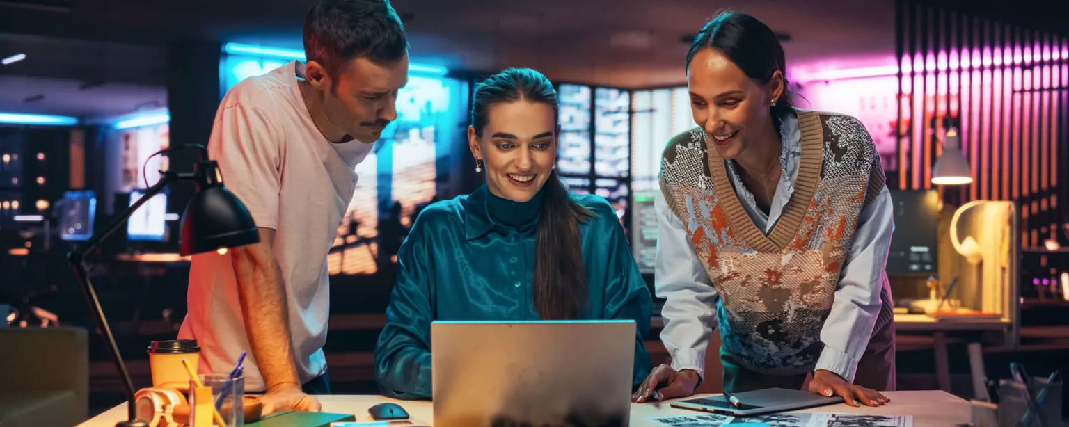 Three marketing professionals collaborate around a laptop in an office equipped with digital marketing tools.